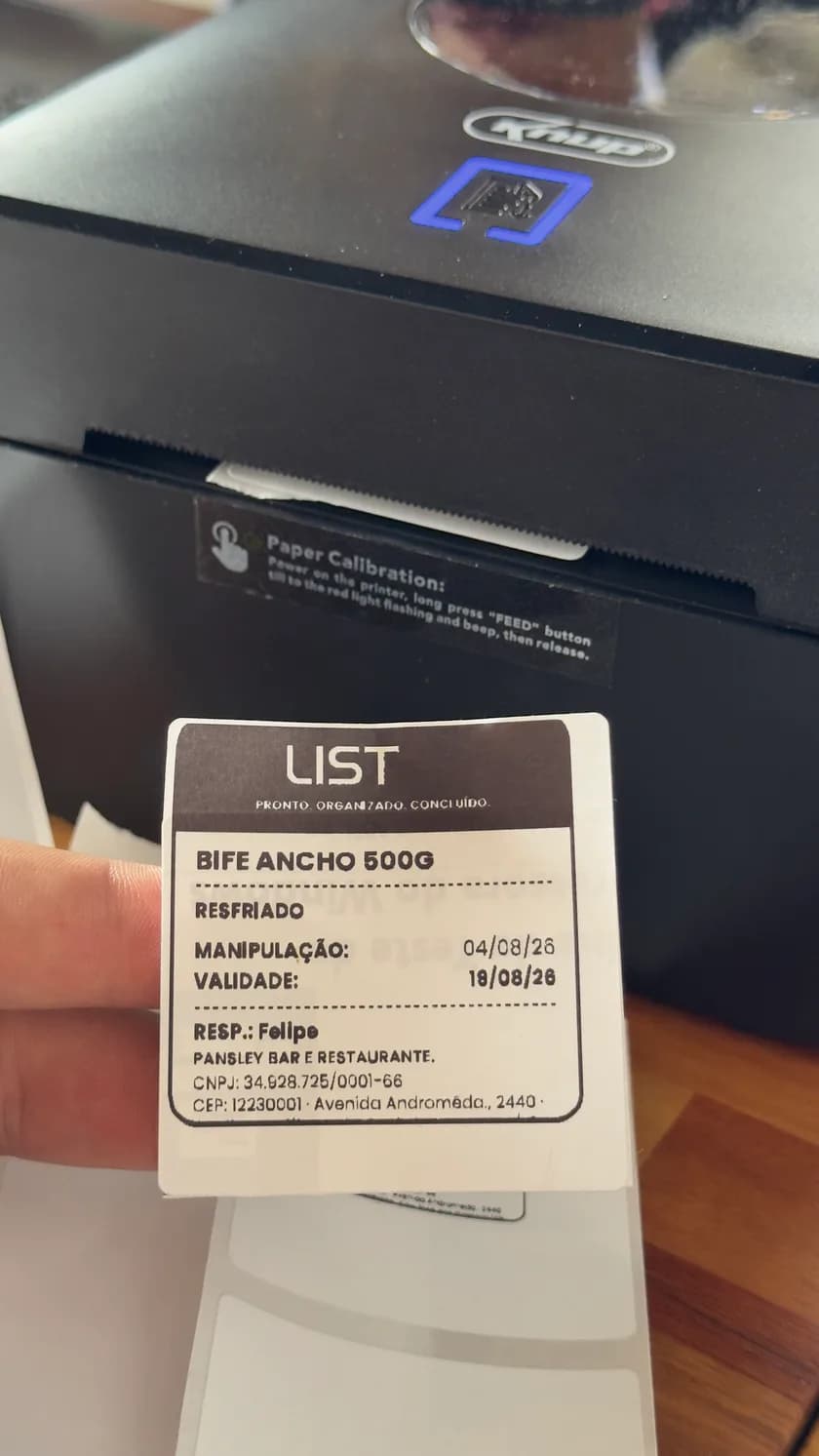 Etiqueta impressa saindo da impressora térmica, com nome do produto, condição de conservação, data de manipulação, data de validade e o responsável.